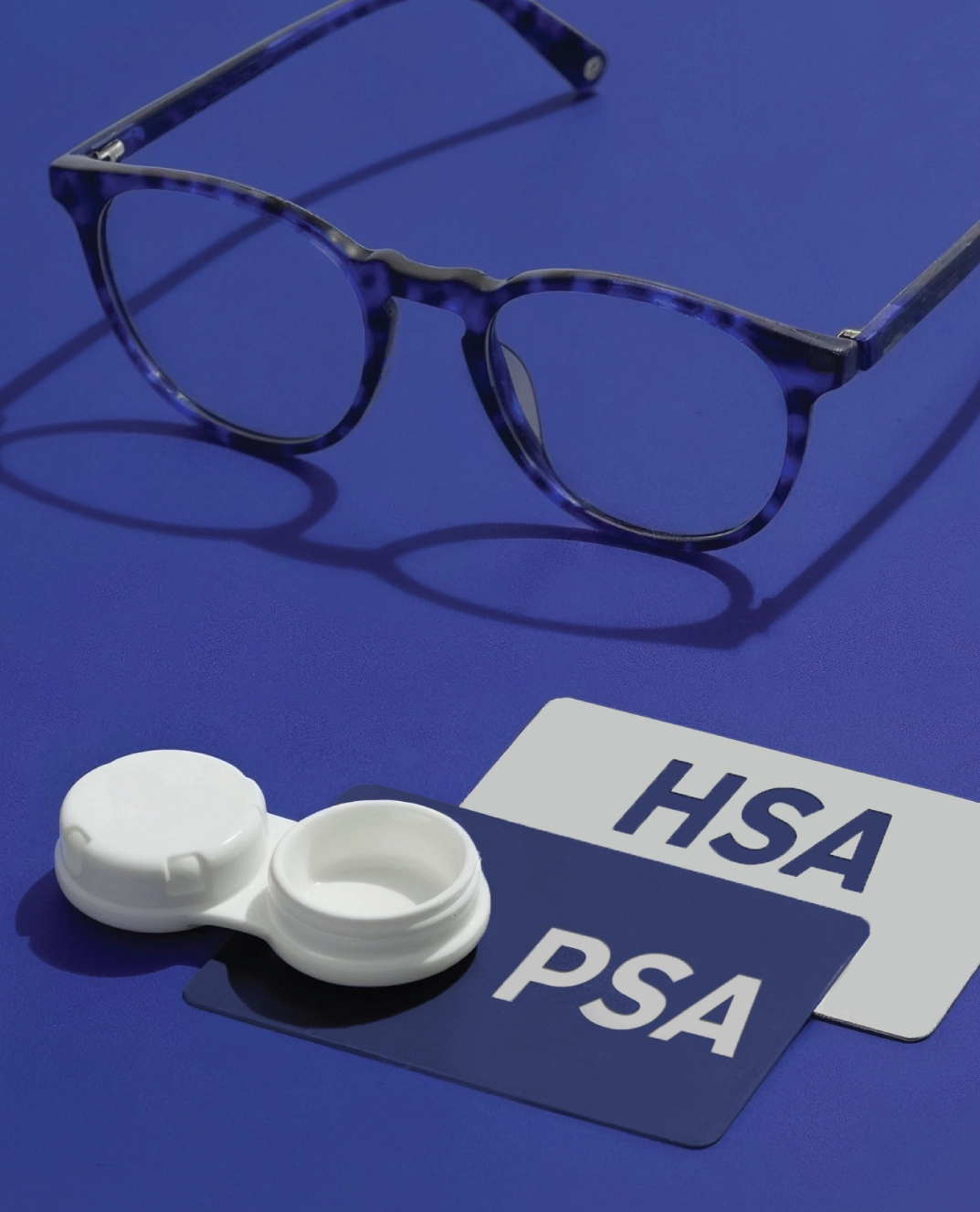 Round eyeglasses with a contact lens case beside FSA and HSA cards on a blue surface, representing eligible vision purchases.