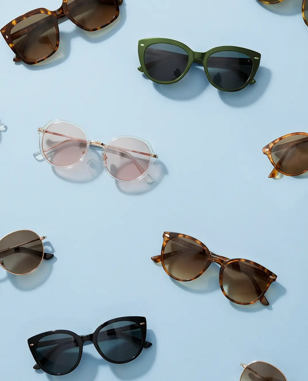 Flat lay of women’s sunglasses — tortoise, black, green, and clear frames — on a light blue background, showcasing trending shapes and styles.