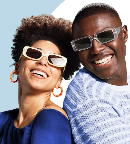 Two friends laughing outdoors while wearing trendy sunglasses and bright clothing.